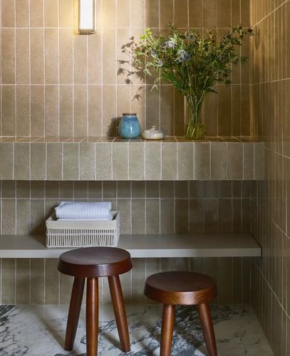 Tiled bathroom niche with floating stone shelves, wooden stools, and a vase of flowers under sconce light