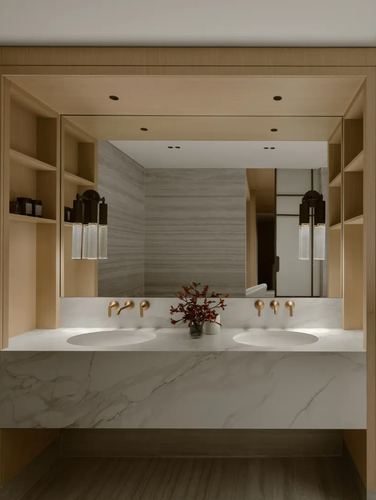 Double vanity with waterfall marble countertop, brass wall taps, and wood-framed mirror niche with open shelving.