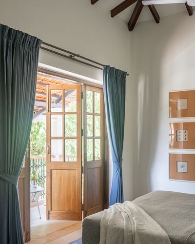 Bedroom with sage curtains framing open wooden french doors leading to a leafy Goan balcony