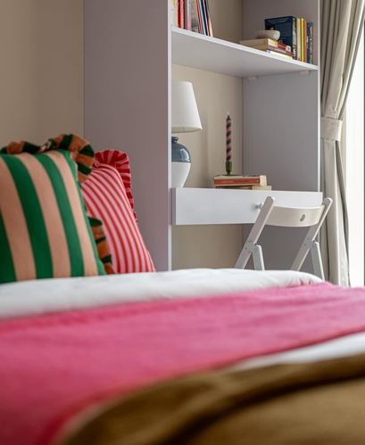 Close-up of a bed with striped pillows and pink throw beside a white built-in shelf and lamp