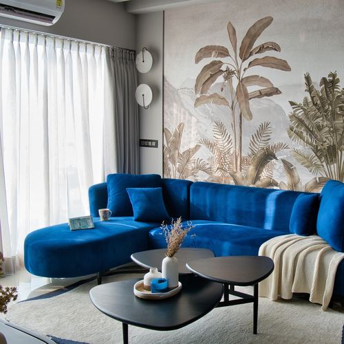 Curved blue velvet sofa in living room with tropical leaf mural and black nesting tables