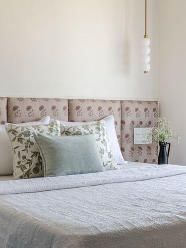 Bed with block-print pink headboard panels, layered cushions, and a bubble pendant light beside a small vase.