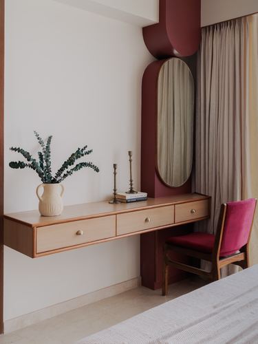 Floating wooden dressing table with mirror set in a maroon arched niche, beside a pink velvet chair