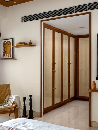 Bedroom corner with teak and cane wardrobe, floating shelf with folk art, cane chair, marble floor