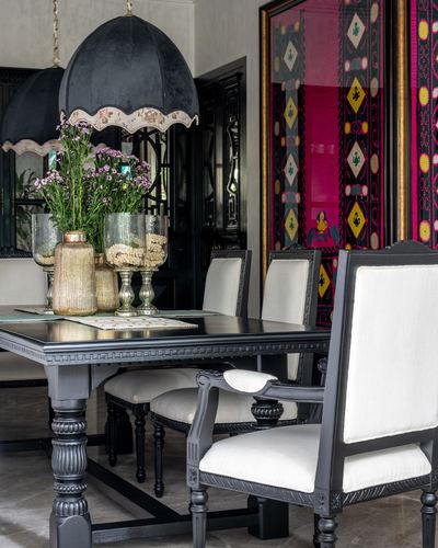 Black carved dining table with ivory chairs beneath scalloped pendant lamps, framed embroidered textile behind