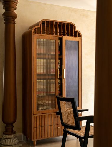 Wooden arched display cabinet with fluted glass doors beside a turned column and cane chair