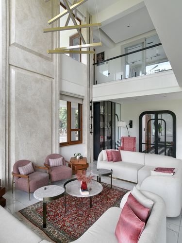 Double-height living room with marble wall, white sofa, pink armchairs and patterned rug below a mezzanine