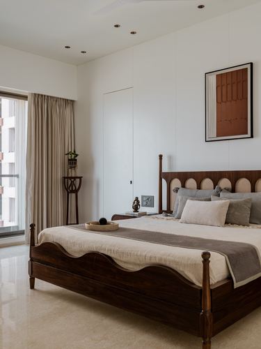 Bedroom with carved wooden bed, neutral linens, and framed terracotta artwork beside balcony curtains