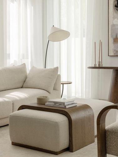 Cream sofa and ottoman with curved wood tray table under sheer curtains and arc lamp
