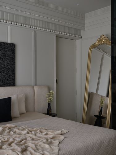 Bedroom corner with beige boucle headboard, gilt mirror, and panelled walls with cornice moulding