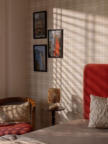 Sunlit bedroom corner with plaid wallpaper, framed photos, rattan lamp, and red velvet headboard