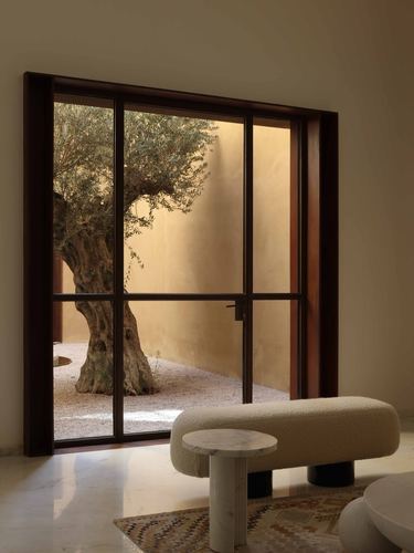 Living room with steel-framed window overlooking a courtyard olive tree, boucle bench and marble table in foreground