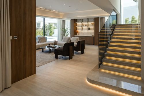 Open-plan living space with wood-panelled wall, glass-railed staircase with amber lighting, and lounge seating beyond