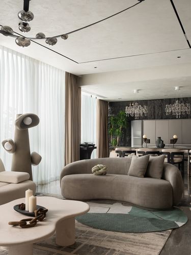 Curved beige sofa with green cushion faces round rug in living room with sheer curtains and dark textured wall beyond
