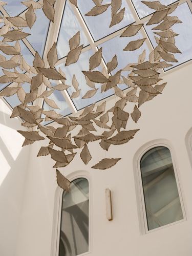 Leaf-shaped hanging sculpture beneath a triangular glass skylight above arched windows in a white stairwell