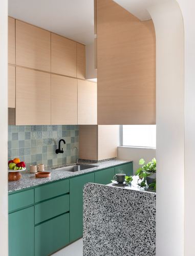 Kitchen with wood upper cabinets, teal lower cabinets, terrazzo counters and island, seen through curved archway