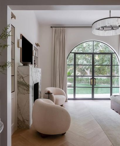 Living nook with veined marble fireplace, boucle chairs, and arched glass doors overlooking a pool and trees