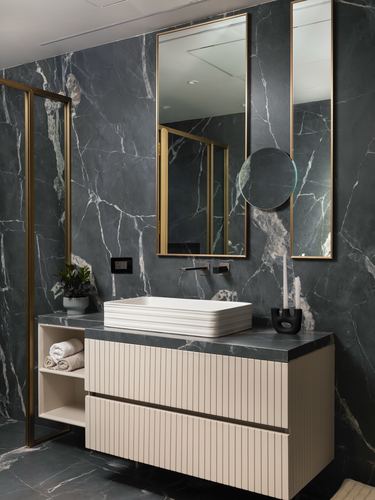 Bathroom vanity with fluted cream cabinetry, brass mirrors, and dark veined marble walls