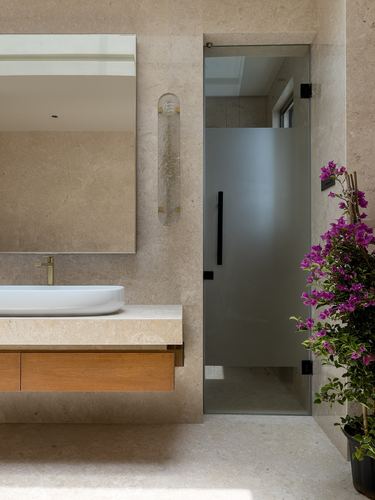Stone-clad bathroom with floating wood vanity, mirror, and frosted glass shower door beside flowering plant