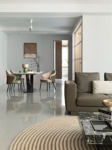 Open-plan living and dining space with taupe sofa, marble coffee table, and dining table beneath an abstract painting, reflected on glossy floor.