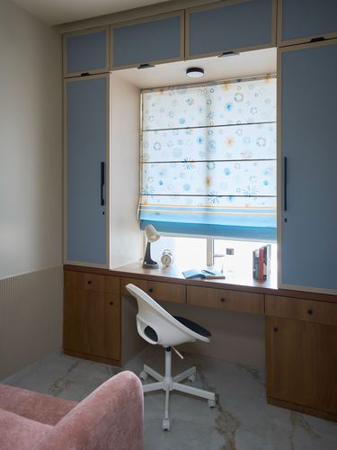 Study nook with wooden desk under window, patterned blind, blue built-in cupboards, pink armchair in foreground
