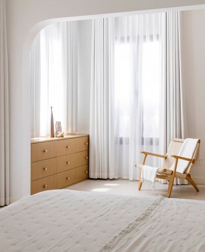 Bedroom corner with arched niche, sheer curtains, wooden dresser, and woven lounge chair by window