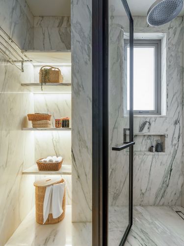 Marble bathroom with backlit shelves holding rattan baskets beside a glass-enclosed shower.