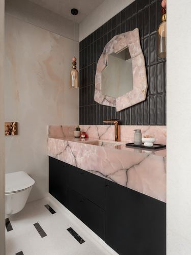 Pink stone vanity with black cabinetry beneath a hexagonal stone-framed mirror on ribbed black tile wall.