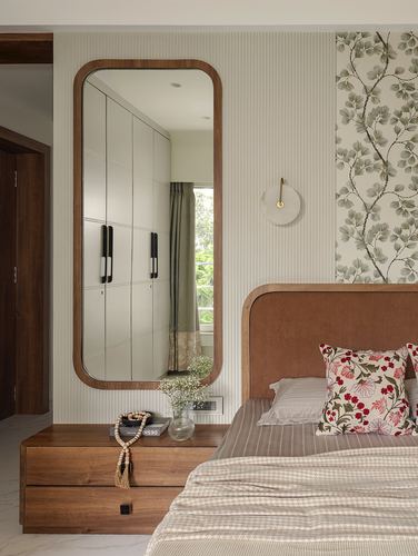 Bedroom corner with tall wood-framed mirror reflecting wardrobe, floral wallpaper accent wall, upholstered headboard