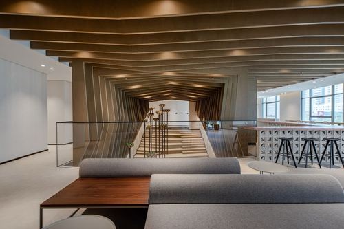 Office lounge with ribbed wood ceiling framing a staircase void and glass railing, grey sofas in foreground