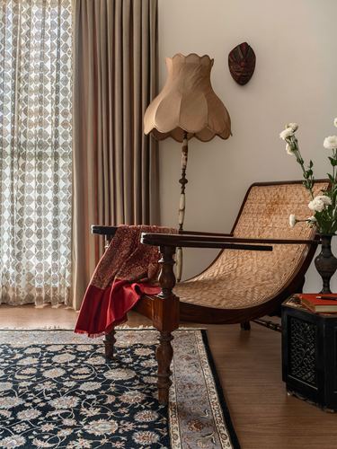 Caned curved lounge chair beside a scalloped lamp, carved table with shawl, patterned rug on wood floor