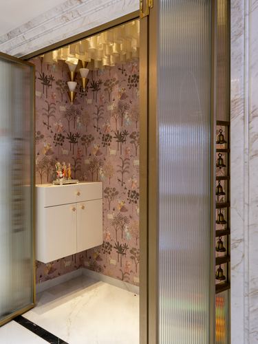 Pooja niche with pink patterned wallpaper, white cabinet, brass bells and fluted glass door in marble hallway