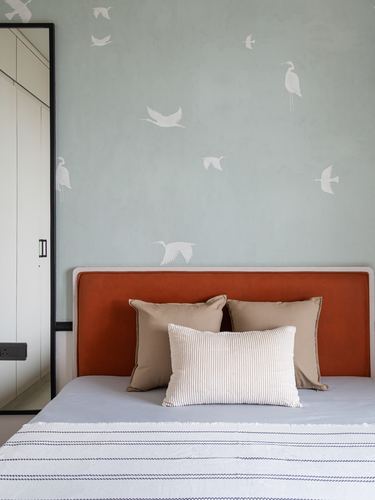 Bed with orange velvet headboard against sage wall painted with white cranes, wardrobe at left
