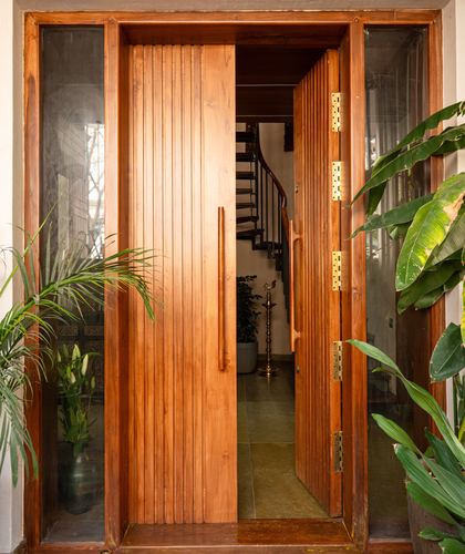 Fluted wooden front door open to reveal spiral staircase and stone floor foyer, flanked by plants