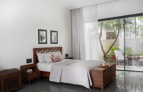Bedroom with carved wood bed, trunk bench, and sheer curtains opening to a green courtyard