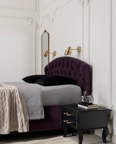 Plum velvet tufted headboard bed against white molded wall with brass sconces and black nightstand