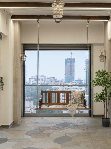 Wooden swing hangs before a large window framing Mumbai skyline, on a stone-tiled balcony.