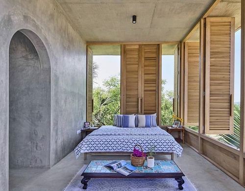 Bedroom with concrete walls, wooden louvered shutters, and blue patterned bedding overlooking greenery