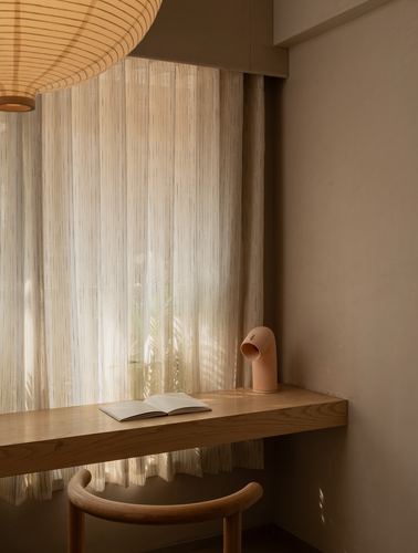 Wooden window desk with open book and ceramic sculpture beneath sheer curtains and a woven pendant lamp