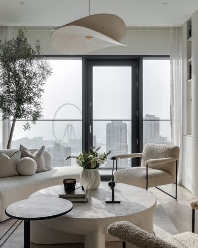 Neutral toned living room with boucle armchairs, stone coffee table, olive tree, and floor-to-ceiling city view windows