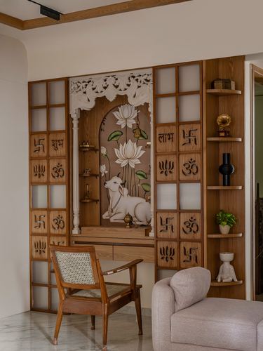 Wooden pooja unit with carved swastik panels, painted cow mural, cane chair and boucle sofa in foreground