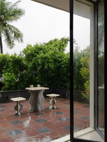 Terrace with curved stone table and stools facing green hedge, seen through open glass door.