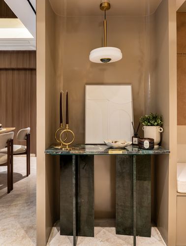 Taupe niche with green stone console table, brass pendant light, and framed artwork beside a dining area