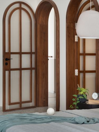 Arched teak doors with fluted glass panels open into a bedroom with terrazzo flooring and pendant light