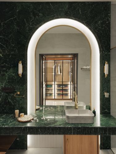 Green marble bathroom vanity with arched illuminated mirror reflecting a walk-in closet beyond