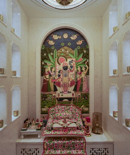 Marble pooja room with arched niche holding a pichwai painting above floral-upholstered deity seating and brass diyas.
