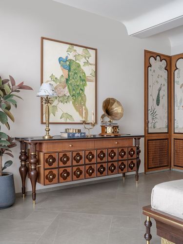 Carved wooden sideboard with brass fittings below a framed peacock painting, beside a floral folding screen