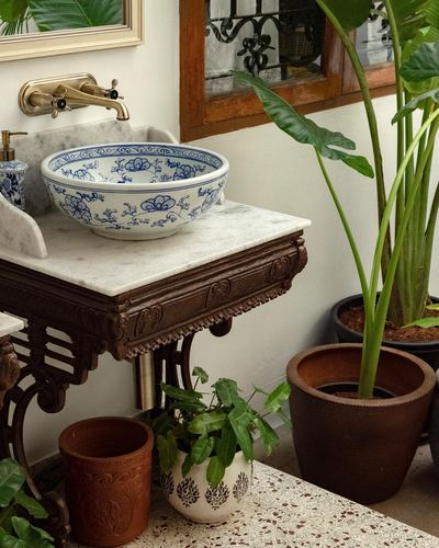 Blue and white ceramic vessel sink on marble-topped carved wooden vanity with brass faucet and surrounding potted plants