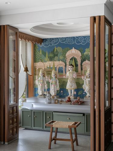 Pooja room with marble deity idols against a painted mural, green cabinetry below, wooden doors