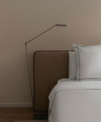Minimalist bedroom corner with taupe upholstered headboard, white bedding, and an angular metal floor lamp against a neutral wall.
